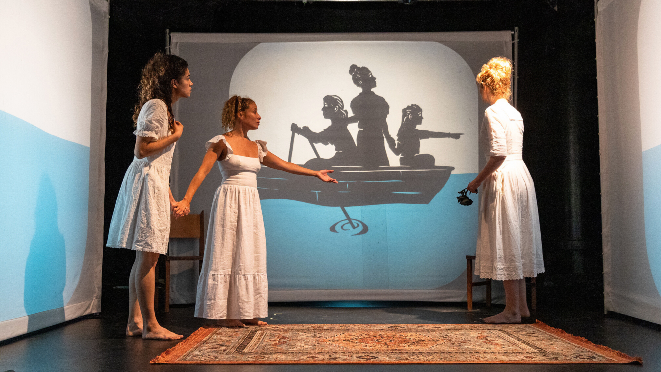Creative Opportunity Fund. Three women in white dresses stand on stage looking at a projected shadow image of people in a boat.